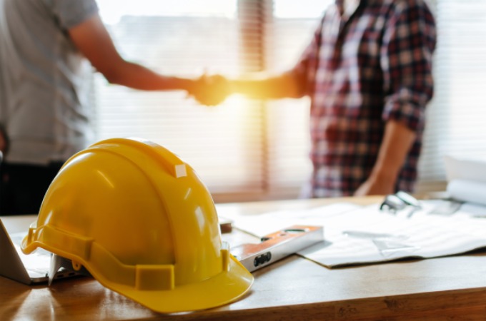 Construction helmet over house blue prints with two man shaking hands in the background