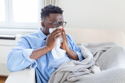 Ill african young woman covered with blanket blowing running nose got fever caught cold sneezing in tissue sit on sofa, sick allergic black girl having allergy symptoms coughing at home, flu concept