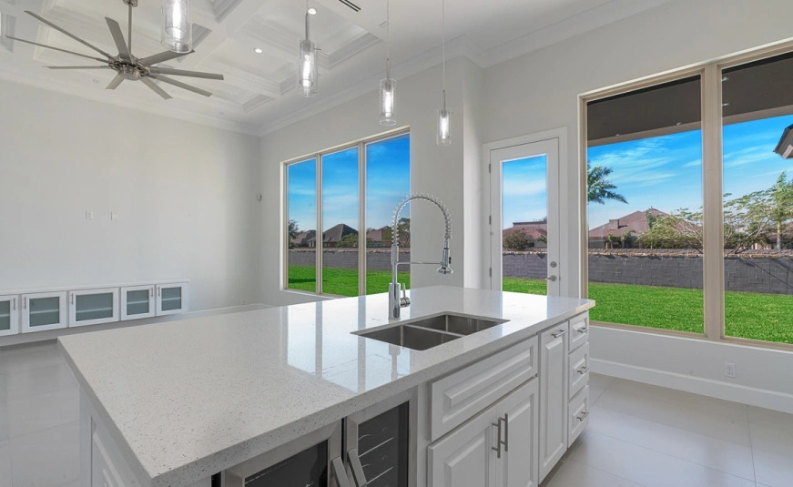 Modern kitchen with a large island, stainless steel sink, and large windows showcasing a green backyard and blue sky.