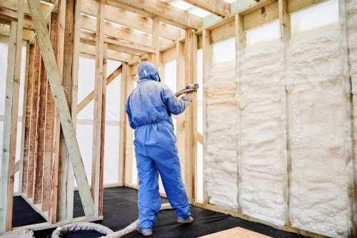 Worker spraying polyurethane foam insulation into timber wall cavities inside unfinished house. Interior construction scene, thermal sealing, weatherization and energy efficient building concept
