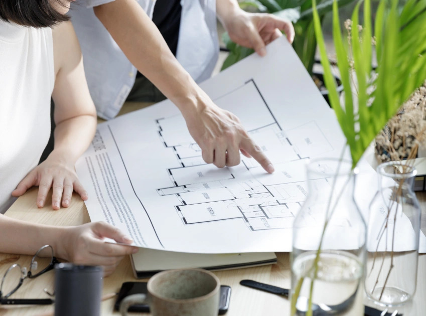 Two people examining a floor plan on a table, with plants and office supplies in the background.